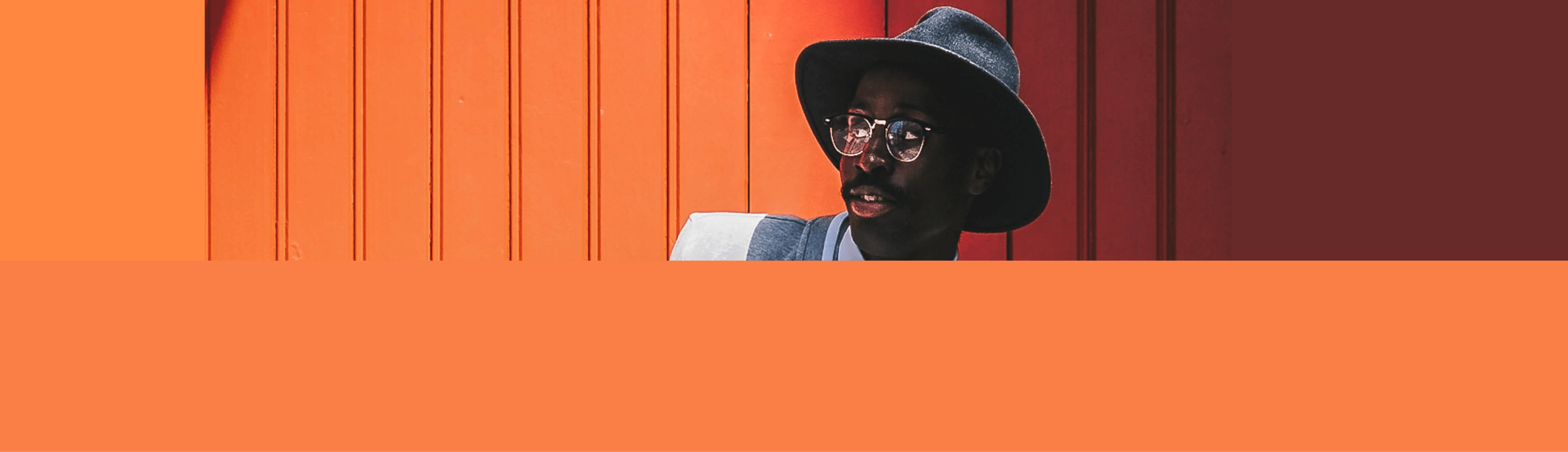 mModelo masculino vestindo chapéu preto, blazer cinza, camisa social branca, gravata borboleta na cor preta, óculos com lente transparente e armação na cor preta, além de um cordão prateado, em um fundo laranja