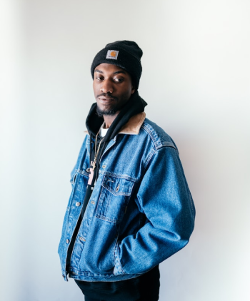 Modelo masculino vestindo toca e calça na cor preta, jaqueta jeans azul, blusa branca e cordão dourado, em um fundo branco