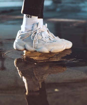 Foco nos pés de modelo usando par de tênis e meia na cor branca, e calça na cor preta, pisando em uma poça de água