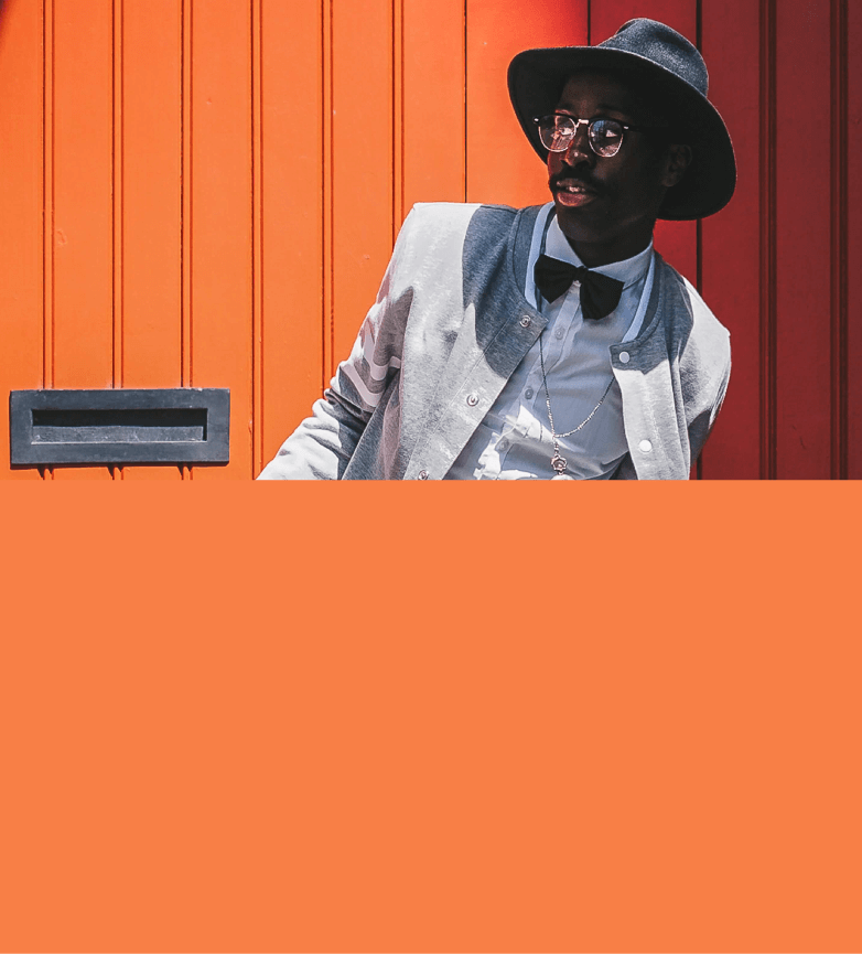 Modelo masculino vestindo chapéu preto, blazer cinza, camisa social branca, gravata borboleta na cor preta, óculos com lente transparente e armação na cor preta, além de um cordão prateado, em um fundo laranja