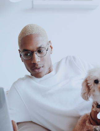 Modelo masculino com blusa branca e um par de óculos de lentes transparentes arredondadas, utilizando um notebook, com um cachorro poodle branco no seu colo, e em um fundo branco