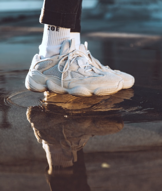 Foco nos pés de modelo usando par de tênis e meia na cor branca, e calça na cor preta, pisando em uma poça de água